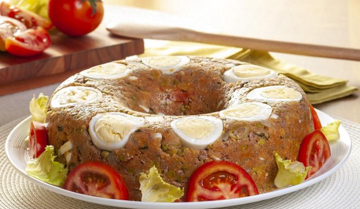 Fotografia em tons de amarelo em uma bancada de madeira de cor branca. Ao centro, um prato branco contendo o cuscuz e ao fundo, há uma tábua de madeira com tomates e uma colher de pau.