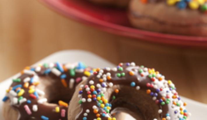 Fotografia em tons de marrom, branco e vermelho de uma bancada de madeira, sobre ela um prato branco com um donuts. Ao fundo um prato vermelho com donuts.