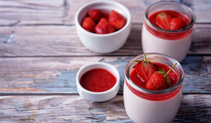 Fotografia de dois recipientes de vidro pequenos com mousse e uma calda de morangos e frutas vermelhas, sobre uma mesa de madeira. Ao redor tem dois recipientes, com morangos inteiros e o outro, com um pouco da calda.