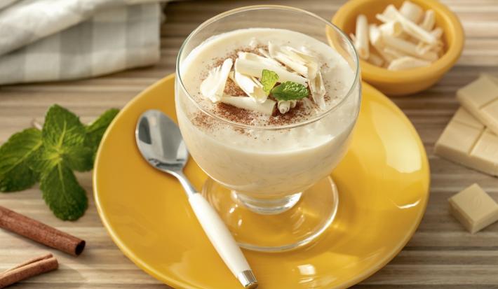 Fotografia em tons de branco em uma bancada de madeira de cor marrom. Ao centro, um pires amarelo contendo uma taça com o arroz doce. Ao lado, há uma colher e alguns pedaços de chocolate, canela e folha de hortelã. Ao fundo, um pano quadriculado cinza.