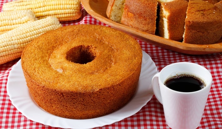 Fotografia de um bolo de milho com coco, exibido em um prato branco sobre uma toalha de mesa xadrez vermelha e branca. O bolo, com crosta dourada, é acompanhado por espigas de milho e uma caneca de café.