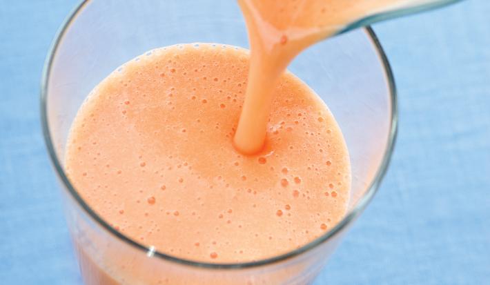 fotografia em tons de azul e laranja vista de uma bancada azul, contém um copo transparente com a vitamina e uma jarra despejando a vitamina no copo