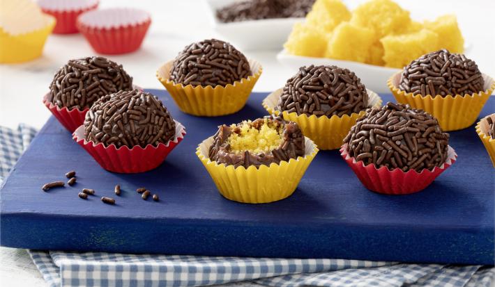 Fotografia em tons de azul, vermelho, amarelo e marrom de uma bancada com um pano quadriculado azul e branco, uma tábua azul, com 7 brigadeiros em cima. Ao centro um brigadeiro mordido, mostrando o recheio de bolo de cenoura.