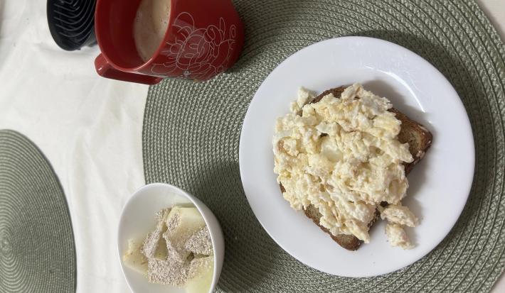 Fotografia de um prato raso médio branco com uma fatia de pão e ovos mexidos cremosos com farelo de aveia. Ao lado tem um recipiente pequeno de vidro e branco com melão e aveia e uma xícara vermelha grande.
