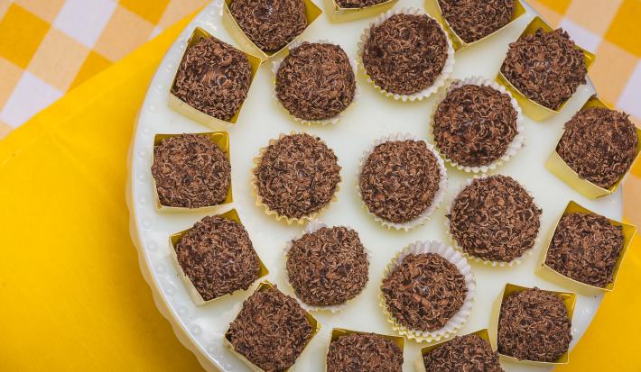 Fotografia em tons de amarelo em uma bancada de madeira com um pano amarelo e outro amarelo xadrez com branco. Em cima deles, um suporte para bolo branco com vários brigadeiros em forminhas amarelas. Fotografia em tons de amarelo em uma bancada de madeira com um pano amarelo e outro amarelo xadrez com branco. Em cima deles, um suporte para bolo branco com vários brigadeiros em forminhas amarelas.