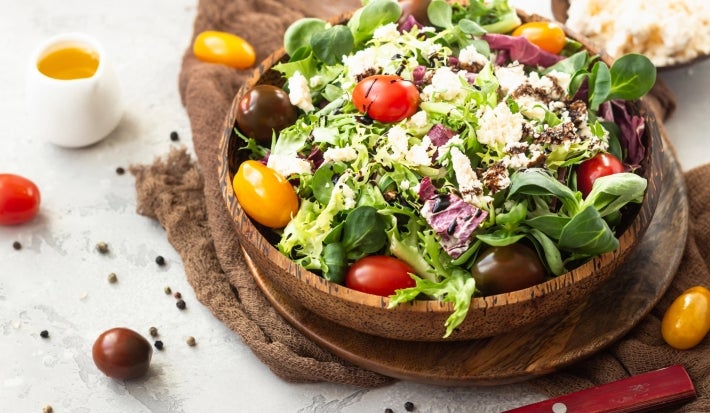 Fotografia de uma salada vibrante em uma tigela, com uma mistura de folhas verdes, tomates-cereja em várias cores e queijo branco esfarelado, regada com molho balsâmico. A composição tem um recipiente de azeite e grãos de pimenta.