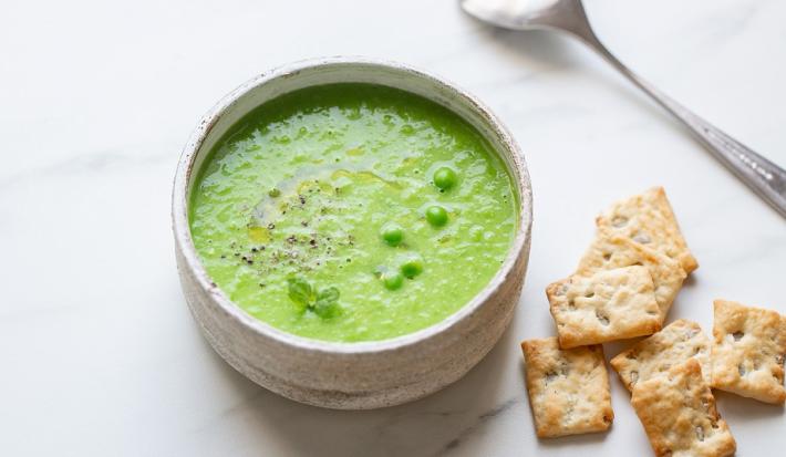 Fotografia em tons de verde em uma bancada de madeira de cor branca. Ao centro, uma tigela contendo a sopa de ervilha com alguns biscoitos de água e sal e uma colher ao lado.