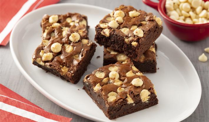 Fotografia em tons de branco, vermelho e cinza de de uma bancada cinza com um prato oval branco, sobre ele cinco pedaços de brownie. Ao fundo um recipiente redondo com gotas de chocolate branco.