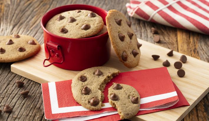 Fotografia em tons de vermelho em uma bancada de madeira com uma tábua de madeira, um guardanapo de papel vermelho com listra branca, um cookie com gotas de chocolate cortado ao meio em cima dele. Ao fundo, uma panelinha vermelha pequena com mais cookies.