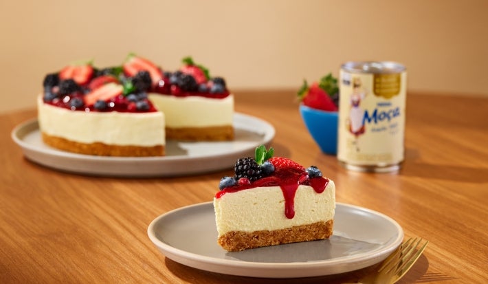 Fotografia mostra uma fatia de cheesecake de frutas vermelha. Ao fundo, o restante da chessecake e a lata de leite moça.