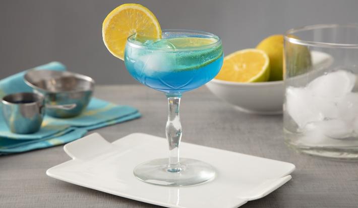 Foto em tons de azul da receita do drink Lagoa Azul servida em uma taça de vidro alta sobre uma base de porcelana branca. Ao fundo há um copo de vidro com cubos de gelo, além de laranjas cortadas e um pano azul para decoração