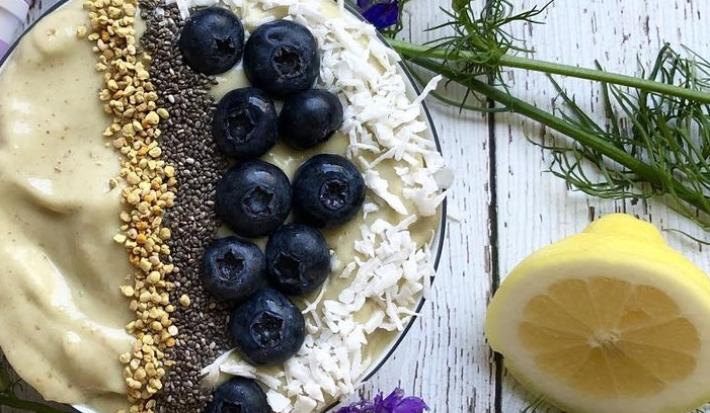 Fotografia em tons de roxo em uma bancada de madeira com um bowl e o creme de avocado dentro dele, decorado com mirtilo, castanha-de-caju e sementes de chia. Ao lado, limão siciliano na mesa.