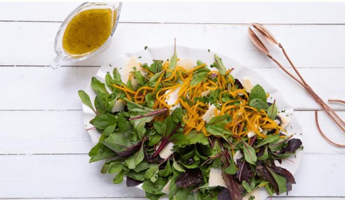 Fotografia em tons de branco e verde, ao centro recipiente com folhas verdes, roxas e tiras de abóbora, ao lado pegador de salada e potinho com molho, tudo sobre bancada na cor branca.