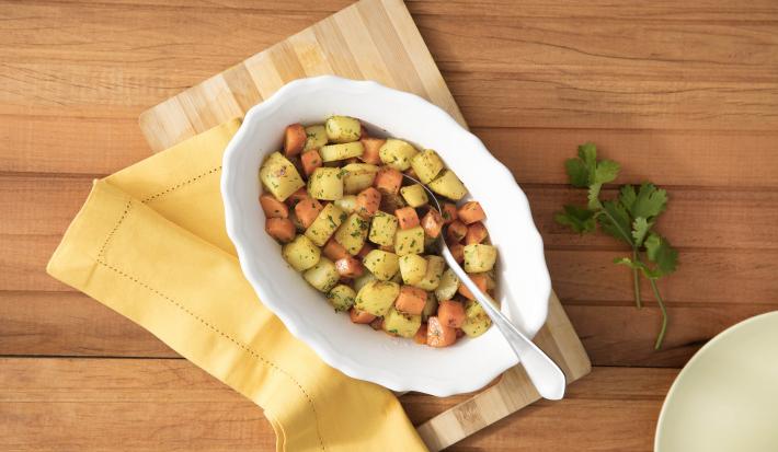 Fotografia em tons de branco, amarelo e marrom, ao centro porcelana branca com legumes cozidos variados polvilhados com coentro picado e uma colher, guardanapo listrado bege e outro amarelo, lado direito prato bege e ramo de coentro, em bancada marrom.