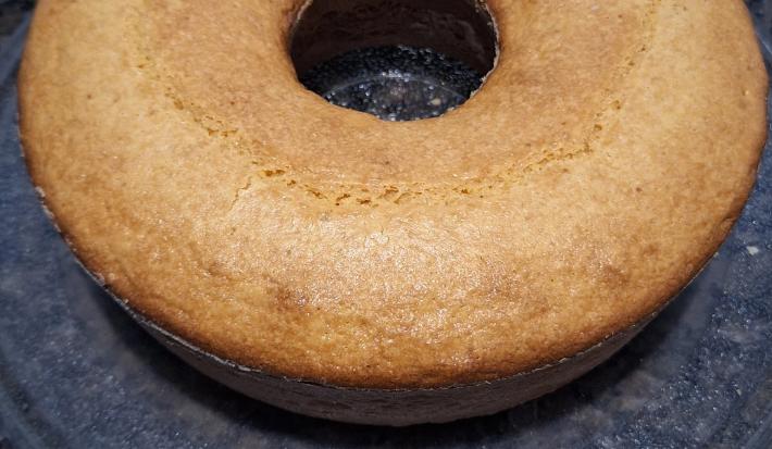 Fotografia de um bolo de fubá, feito na forma de furo central, apoiado em um vidro redondo, sobre uma bancada de cor escura.
