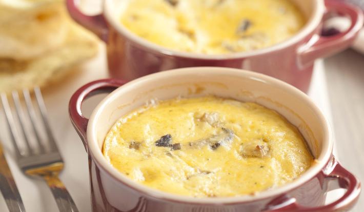 Fotografia em tons de vinho em uma mesa da madeira com uma tábua de madeira bem clara e duas mini panelinhas vinho com o mix de cogumelos gratinado dentro delas. Ao fundo, um pano colorido com detalhes floridos.