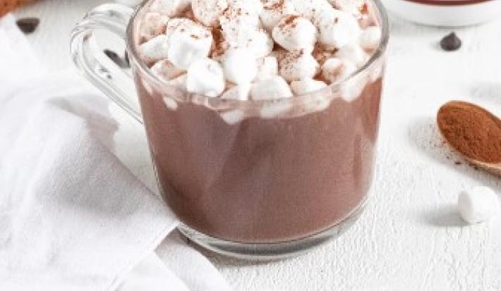 Fotografia em tons de marrom em uma mesa de madeira branca, um pano branco e uma xícara de vidro com o chocolate quente dentro e minis marshmallow para decorar a xícara. Ao lado, uma colherzinha de madeira.