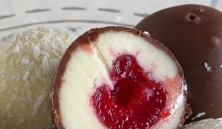Fotografia em tons claros de um bombom de cereja com casquinha de chocolate, tem outro passados apenas no coco ralado.