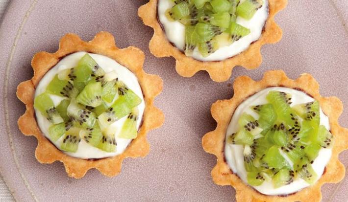 fotografia em tons de lilás, branco e verde tirada de um prato lilás redondo visto de cima, contém 3 mini-torta com pedaços de kiwi e creme.