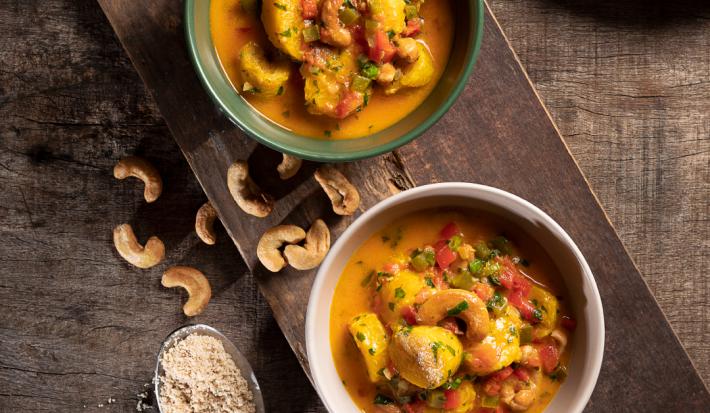 Foto de cima de dois pratos redondos e fundos com a receita de moqueca de banana da terra, ambos sobre uma tábua de madeira, com algumas castanhas-de-caju na mesa, uma colher de metal com arroz e um prato com arroz branco