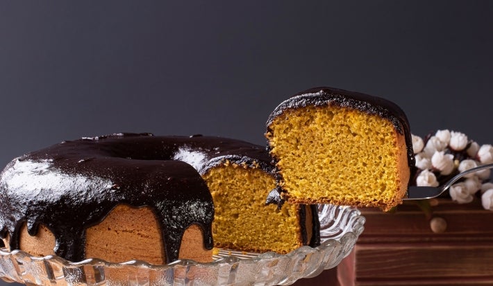 Apresenta o bolo de cenoura inteiro com uma fatia sendo retirada com uma espátula. A cobertura de chocolate está bem brilhante e espessa, escorrendo sobre o bolo. O fundo é escuro e elegante, destacando bem a cor dourada do bolo.