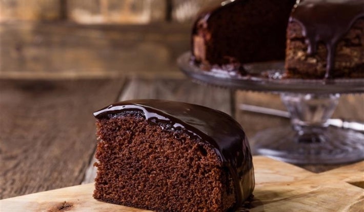 Fotografia de um bolo de chocolate fofinho com uma fatia em destaque, com uma cobertura escura e lisa. Ao fundo, um bolo está em um suporte de vidro, em um ambiente rústico.