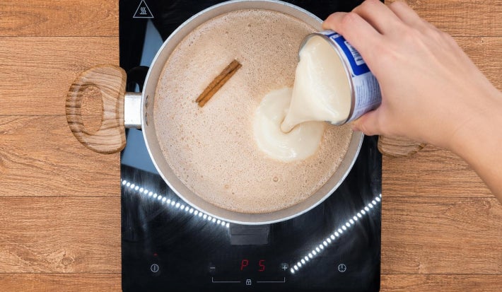 Mão despejando creme de leite sobre panela com ingredientes.
