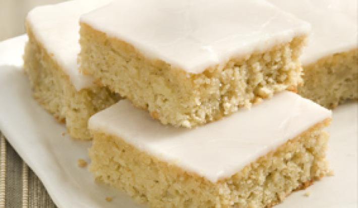 Fotografia em tons de bege em uma mesa de madeira com uma toalha bege listrada, um prato branco quadrado com pedaços de bolo de camomila com cobertura de glacê branco.