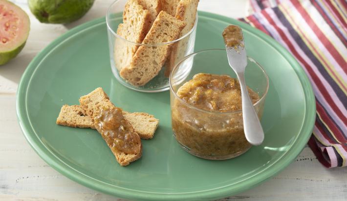 Foto da receita de Geleia de Casca de Goiaba. Observa-se um prato verde com um potinho transparente com a geleia junto com um pote de torradinhas para acompanhar