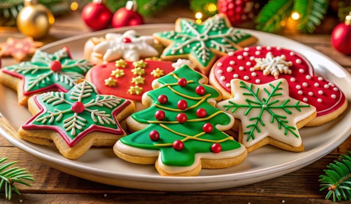 Em cima de uma mesa, prato com biscoitos em formato de árvores de Natal, estrelas, decorados com glacê colorido e confeitos coloridos de bolinhas vermelho.