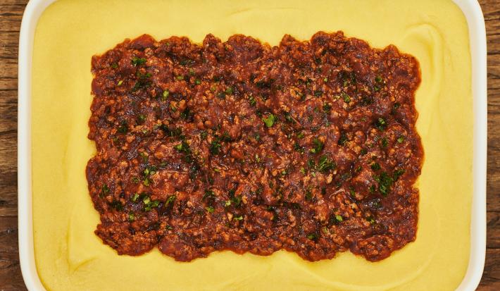 Foto bem aproximada de uma travessa branca. Dentro dela está a receita de polenta em tom amarelo claro e, ao centro, a carne moída com molho