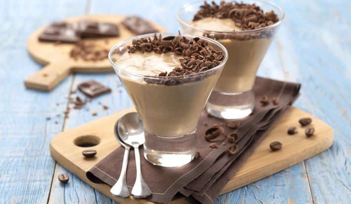 Foto da receita de mousse de cappuccino. Observa-se uma superfície de madeira azul-clara, em cima dela uma pequena tábua de madeira com um guardanapo marrom e duas taças de vidro com a mousse apoiadas. Como decoração, raspas de chocolate e grãos de café