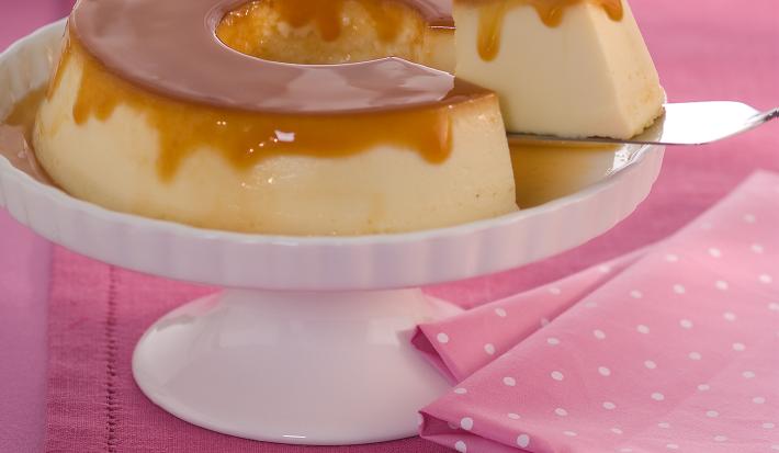 Foto de uma bancada rosa, com um tecido rosa de bolinhas brancas. Ao centro há um prato branco alto com um Pudim com calda de caramelo e um pedaço dele está sendo retirado com uma espátula. Ao fundo há 3 pratos brancos empilhados.