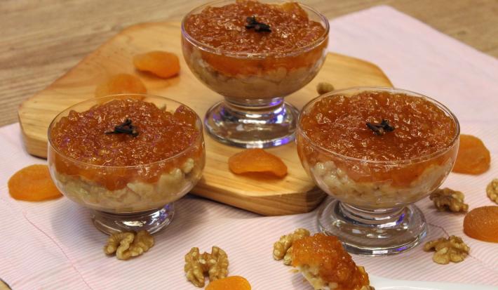 Fotografia em tons de laranja em uma bancada de madeira de cor marrom. Ao centro, uma toalha rosa com uma tábua de madeira. Em cima, estão as taças de brigadeiro e ao redor, damascos.