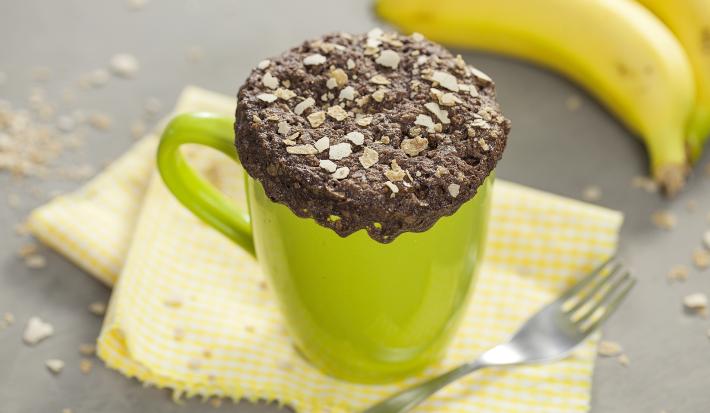 fotografia em tons de cinza, verde e marrom de uma bancada cinza vista de cima. Contém um pano amarelo, uma xicara verde com um bolo de chocolate com cereais por cima e um garfo ao lado e um cacho de bananas