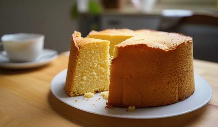 Um bolo grande e fofo de cor amarela clara, com uma fatia já cortada. Ele está sobre um prato branco, em uma mesa de madeira clara. Ao fundo, há uma xícara branca desfocada, sugerindo um ambiente aconchegante de café da manhã ou lanche da tarde.