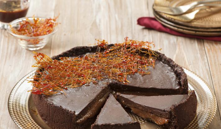 Fotografia em tons de marrom e chocolate, mostrando bancada em tons de marrom e branco contendo prato dourado com uma torta de chocolate decorada com fios de caramelo, ao fundo pratos e garfos, potinho com calda de caramelo e potinho com fios de caramelo.
