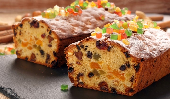 Fotografia de um bolo de frutas natalino, com uma camada generosa de frutas cristalizadas coloridas no topo. O bolo, cortado, revela um interior rico e vibrante.