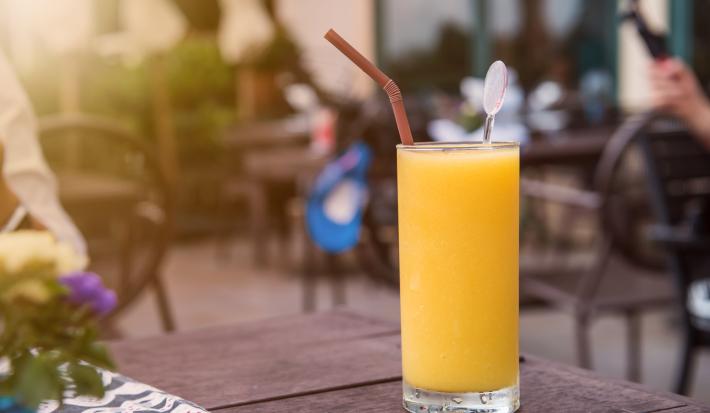 Fotografia de um copo grande com um refresco de laranja, dentro do copo, um canudo de cor marrom e um enfeito de vidro. O copo está sobre uma mesa de madeira na cor marrom. Ao fundo, mesas e cadeiras de uma cafeteria ao ar livre.