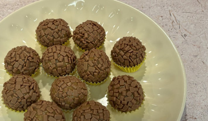 Docinhos cobertos com granulado de chocolate em prato claro, com latas de Leite Moça ao fundo.