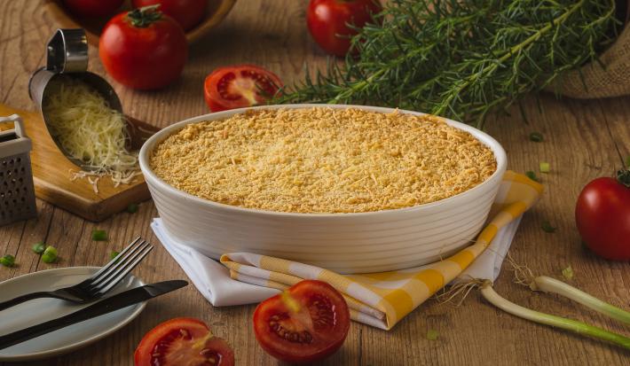 Fotografia em tons de amarelo em uma bancada de madeira escura, um recipiente branco oval com o escondidinho de carne suína gratinado dentro dele, um pano amarelo xadrez e alguns elementos para compor a foto, como tomate, queijo ralado e alecrim.