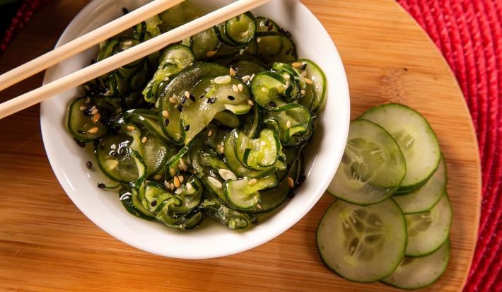 Imagem de um bowl branco sobre uma tábua de madeira com a salada de pepino em fatias e gergelim com um hashi.