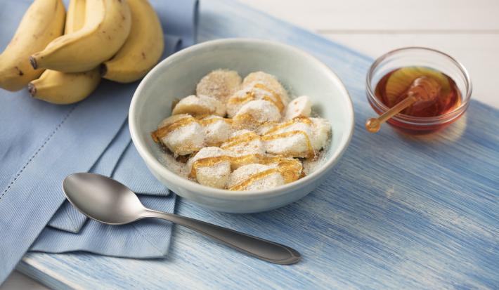 Fotografia em tons de azul em uma mesa de madeira clara com um paninho azul ao lado, um prato branco redondo fundo com as rodelas de banana cobertas com mel, aveia, leite em pó e linhaça. Ao lado, um cacho de bananas e um potinho com mel.