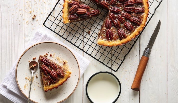 Fotografia vista de cima em tons claros de uma fatia de torta de nozes e doce de leite em um prato raso médio branco. Ao lado tem o restante da torta sobre uma grade preta.