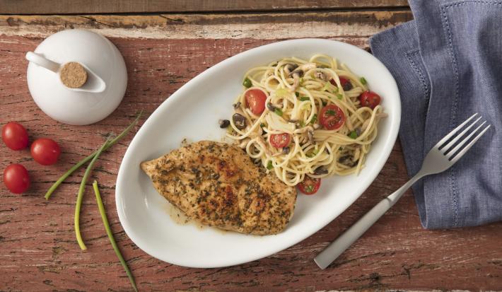 Fotografia em tons de azul e vermelho em uma bancada de madeira com um pano azul ao lado, um recipiente oval branco fundo com o macarrão com cogumelos e tomates e ao lado um filé de frango.