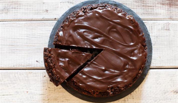 Fotografia em tons de marrom e branco de uma bancada marrom vista de cima, contém um prato redondo e branco com um bolo de chocolate com cobertura de chocolate e uma fatia sendo retirada por uma espátula