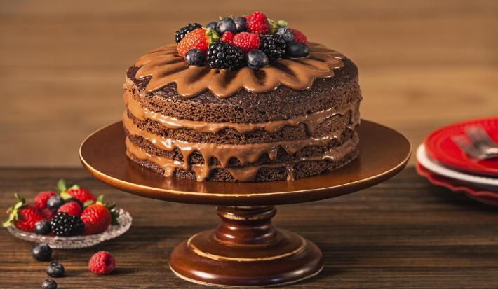 Fotografia em tons de marrom em uma bancada de madeira escura com um suporte para bolo marrom e o naked cake com ganache de cacau e frutas vermelhas em cima. Ao lado, um pratinho de vidro com frutas vermelhas.