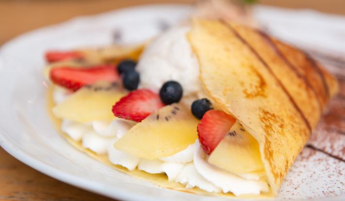 Fotografia em tons de branco com um prato branco ao centro. Em cima do prato existe um crepe recheado com um creme branco doce, morango, kiwi e blueberries.