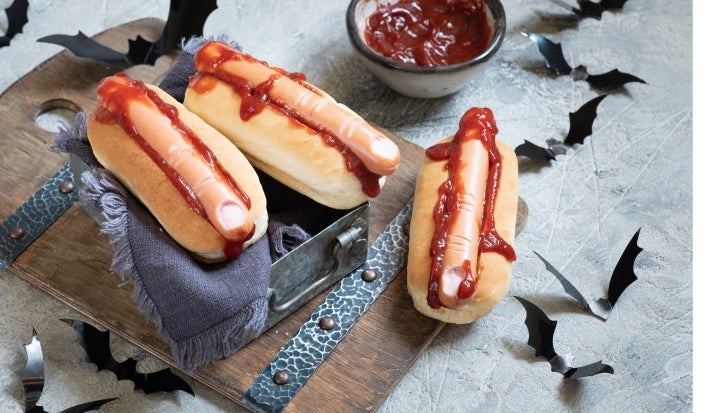 Cachorro-quente com salsichas em formato de dedo, pão macio e dourado, corte superficial na ponta simulando unha, com ketchup vermelho escorrendo.