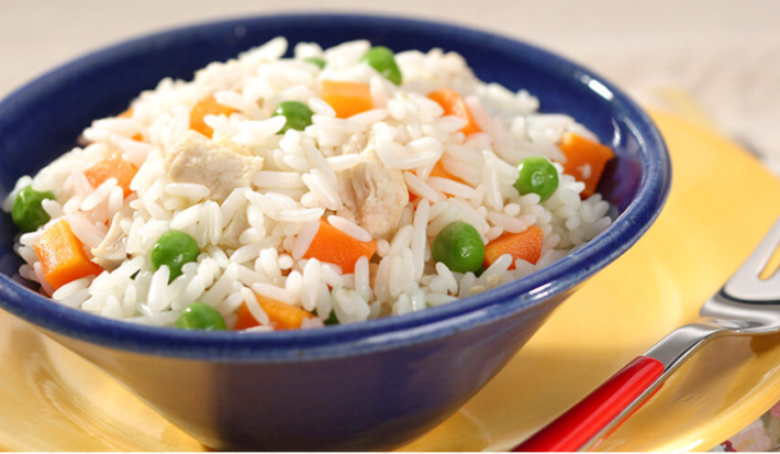 fotografia em tons de amarelo, azul e branco tirada de um prato redondo amarelo com um recipiente azul, que contém o arroz com pedaços de cenouras e ervilhas com um garfo ao lado para servir